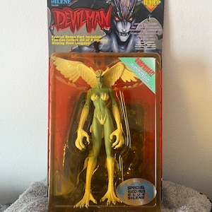 May include: A plastic action figure of a green-skinned female demon with wings and bird-like feet. The figure is packaged in a clear plastic blister pack with a red and yellow background. The packaging features the text "Devilman" and "Silene".