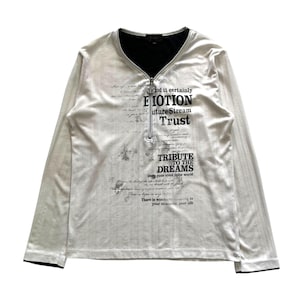 Op de afbeelding: Een wit shirt met lange mouwen, een zwarte V-hals en een kwartrits. Het shirt heeft bedrukte tekst, waaronder de woorden "Emotion", "Trust" en "Tribute to the Dreams."