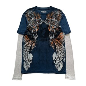 Könnte beinhalten: Ein marineblaues Langarmshirt mit Totenkopf- und Knochenkreuz-Design. Das Shirt hat einen grauen Untergrund und eine Grafik mit zwei geflügelten Totenköpfen mit einem Kreuz und verzierten Details.