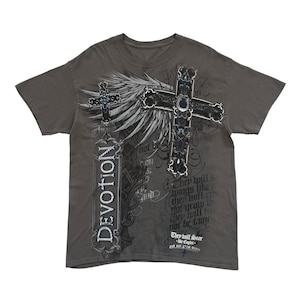 Può includere: T-shirt grigia con un design a croce dettagliato, ali e la parola "DEVOTION". Il design incorpora accenti blu e scritte. La maglietta ha un girocollo e maniche corte.