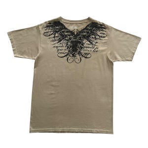 Pode incluir: Camiseta bege de manga curta com um design gráfico preto de asas e escrita. A camiseta tem gola redonda e é feita de um material macio. O gráfico é centrado no peito.