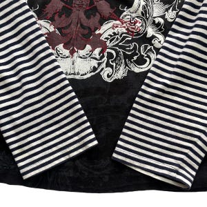 May include: Black long-sleeved shirt with a large ornate design in burgundy and white on the chest. The sleeves are striped with navy blue and white. The shirt has a crew neck and a fitted silhouette.