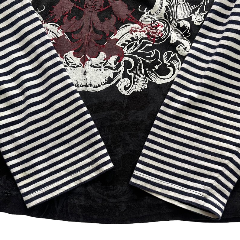 May include: Black long-sleeved shirt with a large ornate design in burgundy and white on the chest. The sleeves are striped with navy blue and white. The shirt has a crew neck and a fitted silhouette.
