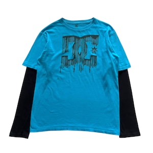 Puede incluir: Una camiseta azul de manga larga con un logotipo DC negro. El logotipo está goteando pintura negra y tiene una estrella encima. La camiseta tiene mangas largas negras que se extienden más allá de la camiseta azul.