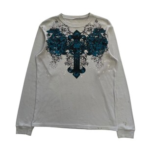 Op de afbeelding: Een langarm, lichtgrijs thermisch shirt met een blauwe en zwarte grafische print van een kruis met sierlijke details.