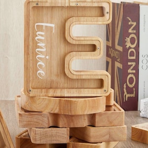 Puede incluir: Una hucha de madera clara con forma de letra E y panel frontal de acr&iacute;lico transparente. El nombre Eunice est&aacute; impreso en letra cursiva blanca en el lado izquierdo del marco de madera. La estructura est&aacute; asegurada con peque&ntilde;os tornillos.