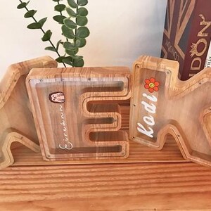 Puede incluir: Tres huchas de madera con forma de letras K, E y K. Cada hucha cuenta con un panel frontal de acr&iacute;lico transparente que permite ver el contenido. Las letras est&aacute;n personalizadas con nombres en letra blanca y peque&ntilde;as pegatinas de colores.