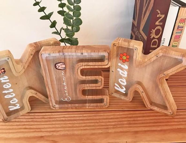 Puede incluir: Tres huchas de madera con forma de letras K, E y K. Cada hucha cuenta con un panel frontal de acr&iacute;lico transparente que permite ver el contenido. Las letras est&aacute;n personalizadas con nombres en letra blanca y peque&ntilde;as pegatinas de colores.