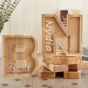 May include: Wooden letter-shaped money boxes. The letter "B" has the name "Basil" in white script, and the letter "N" has the name "Nydia". The money boxes are made of light-coloured wood and are filled with coins. A decorative vase with greenery is in the background.