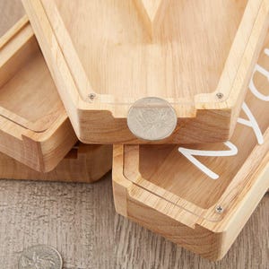 Puede incluir: Una hucha de madera clara con forma de ata&uacute;d y panel frontal de acr&iacute;lico transparente. La madera tiene un acabado de veta natural. Una moneda de color plateado descansa sobre el borde superior del marco. Se ve texto cursivo blanco en el acr&iacute;lico.