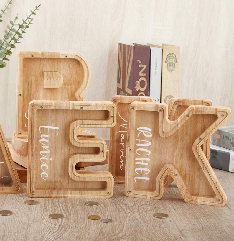 Puede incluir: Una colecci&oacute;n de huchas de madera de color claro con forma de letra y frentes de acr&iacute;lico transparente. Se ven las letras E y K, con los nombres Eunice, Marvin y Rachel impresos en blanco. Hay varias monedas de metal esparcidas cerca.
