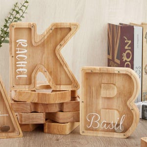 Può includere: Un salvadanaio in legno a forma di lettera K con il nome Rachel stampato in bianco, un salvadanaio a forma di lettera B con il nome Basil stampato in bianco e un salvadanaio triangolare, tutti con frontali in acrilico e viti a vista.