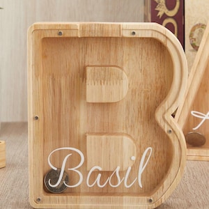 Puede incluir: Una hucha de madera clara con forma de letra B y un panel frontal transparente. El nombre Basil est&aacute; impreso en cursiva blanca en la parte delantera. La estructura se mantiene unida mediante peque&ntilde;os tornillos met&aacute;licos en las esquinas.