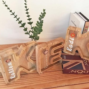 Puede incluir: Tres huchas de madera con forma de letra y frente de acr&iacute;lico transparente. Se ven las letras K, E y K, con los nombres Kolten, Eirinn y Kodi impresos en blanco junto a peque&ntilde;as pegatinas de colores de un mono y una flor.