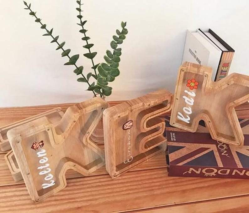 Puede incluir: Tres huchas de madera con forma de letra y frente de acr&iacute;lico transparente. Se ven las letras K, E y K, con los nombres Kolten, Eirinn y Kodi impresos en blanco junto a peque&ntilde;as pegatinas de colores de un mono y una flor.