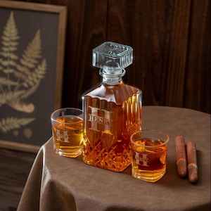 May include: A personalized whiskey decanter set with two glasses and cigars. The amber-colored decanter and glasses are engraved with the name "HENRY". The decanter has a square shape with a diamond pattern. The set is on a brown table.