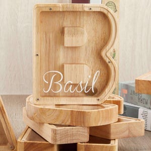 Puede incluir: Una hucha de madera de color claro con forma de letra B y un panel frontal de acr&iacute;lico transparente. El nombre Basil est&aacute; impreso en cursiva blanca sobre la superficie transparente. La hucha muestra tornillos met&aacute;licos y veta de madera.