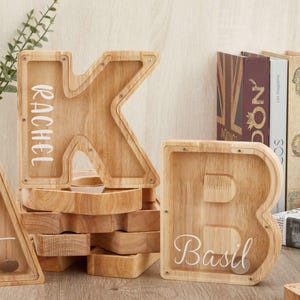 Puede incluir: Una hucha de madera de color claro con forma de letra K con el nombre Rachel impreso en blanco, y una hucha de madera con forma de letra B con el nombre Basil impreso en blanco, ambas con un panel frontal transparente para ver las monedas.