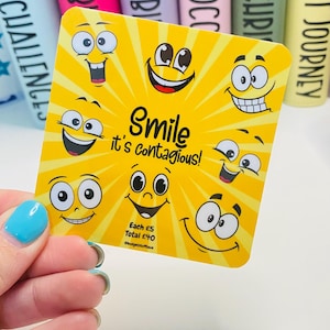 May include: A yellow card featuring various cartoon smiley faces and the text "Smile it's contagious!". The card has a sunburst design. The card is held by a hand with a blue painted fingernail. Colorful books are in the background.