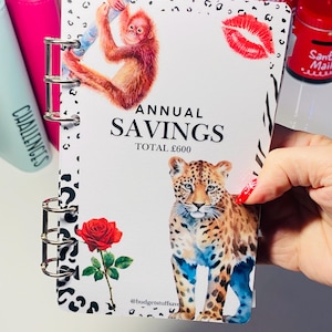 May include: A patterned savings binder with a metal ring binder. The cover features an orangutan, a leopard, a red rose, and a pair of red lips. The text "ANNUAL SAVINGS TOTAL £600" is printed on the cover.