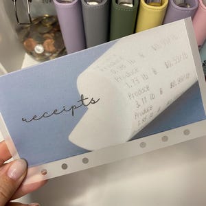 May include: A clear plastic receipt holder with a blue background and the word "receipts" written in cursive. The holder has six holes along the bottom edge and is designed to hold receipts.