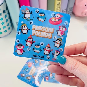 May include: A blue sticker with cartoon penguins in winter clothing. The sticker reads "Penguin Pounds" and "Total £20". The sticker is held up by a hand with blue nail polish. Colourful notebooks are visible in the background.