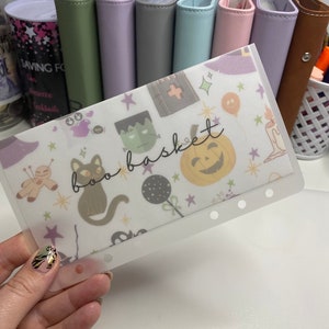 May include: A clear plastic budget envelope with a Halloween-themed pattern featuring a black cat, pumpkin, and the words "boo basket." The envelope is held in front of a row of colorful binders.