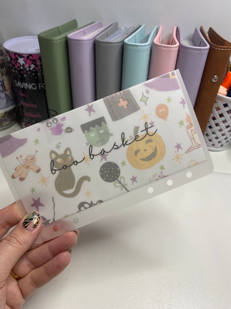 May include: A clear plastic budget envelope with a Halloween-themed pattern featuring a black cat, pumpkin, and the words "boo basket." The envelope is held in front of a row of colorful binders.