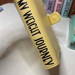 May include: A light yellow planner with the words "MY WEIGHT JOURNEY" printed in black. The planner has a snap closure and is held by a person. Other planners are visible in the background.