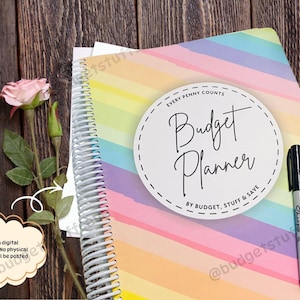 May include: A spiral-bound budget planner with a rainbow striped cover, a black marker, and a pink rose on a wooden surface. The planner has a white circular label that reads "Budget Planner".