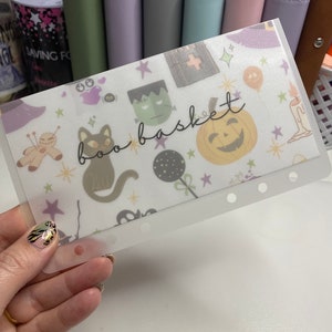 May include: A clear plastic sleeve with a Halloween-themed design. The sleeve features illustrations of a black cat, Frankenstein's monster, a jack-o'-lantern, and the words "boo basket". The background includes stars and other Halloween-related icons.