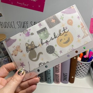 May include: A clear plastic budget envelope with a Halloween-themed pattern. The design includes a black cat, Frankenstein's monster, a jack-o'-lantern, and the words "boo basket". The envelope is held by a hand.