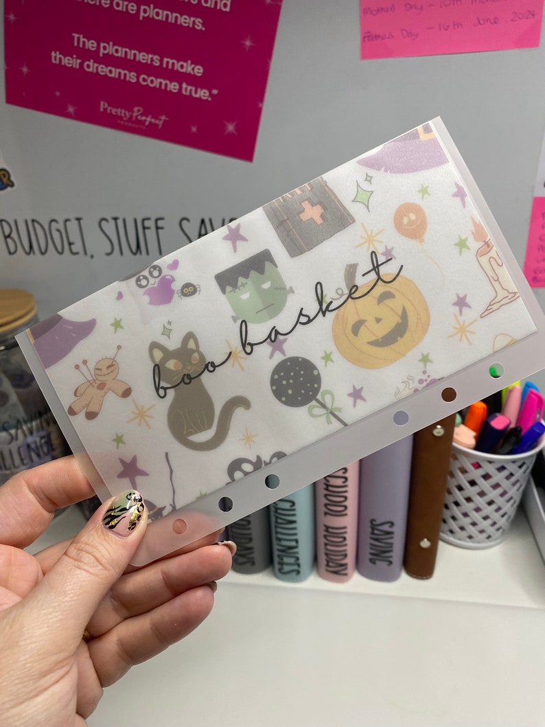 May include: A clear plastic budget envelope with a Halloween-themed pattern. The design includes a black cat, Frankenstein's monster, a jack-o'-lantern, and the words "boo basket". The envelope is held by a hand.