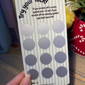 May include: A white card with green stripes and silver circles that can be scratched off. The card says "Try your luck!" and has instructions for a game where you can save £5 or £10. The card also has the text "@budgetstuffsave" at the bottom.
