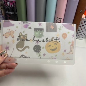 May include: A clear plastic budget envelope with a Halloween-themed design. The envelope features illustrations of a black cat, pumpkin, Frankenstein's monster, and the words "boo basket". The background includes stars and other Halloween icons.