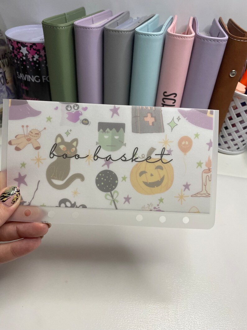 May include: A clear plastic budget envelope with a Halloween-themed design. The envelope features illustrations of a black cat, pumpkin, Frankenstein's monster, and the words "boo basket". The background includes stars and other Halloween icons.