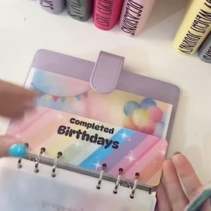 May include: A lilac binder with a tab closure, open to show a rainbow insert labelled "Completed Birthdays." The insert has a balloon graphic and a banner. Other labelled binders are visible in the background.