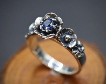 Puede incluir: Un anillo de plata con una piedra preciosa de zafiro azul engastada en un diseño floral. El anillo está adornado con tres flores de plata.