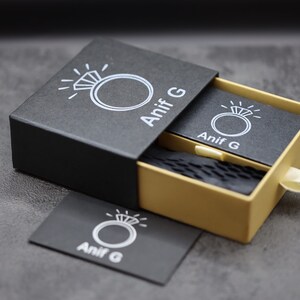 Puede incluir: Una caja de anillo negra con un caj&oacute;n dorado. La caja tiene un logotipo blanco con un anillo y el texto "Anif G".