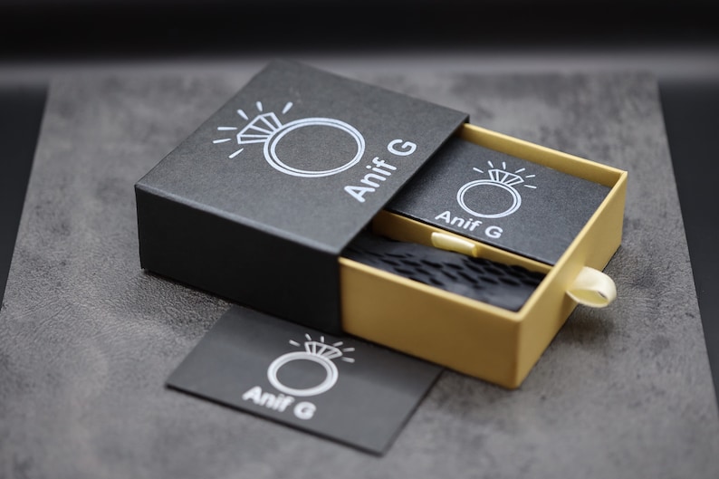 Puede incluir: Una caja de anillo negra con un caj&oacute;n dorado. La caja tiene un logotipo blanco con un anillo y el texto "Anif G".
