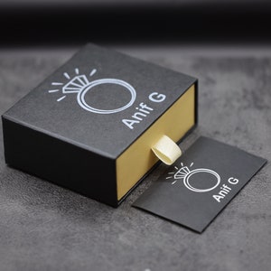 Puede incluir: Una caja de anillo negra con interior dorado y un icono de anillo blanco con el texto "Anif G" en la parte superior. Una peque&ntilde;a tarjeta negra con el mismo icono y texto est&aacute; al lado de la caja.