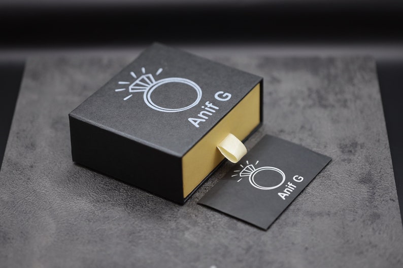 Puede incluir: Una caja de anillo negra con interior dorado y un icono de anillo blanco con el texto "Anif G" en la parte superior. Una peque&ntilde;a tarjeta negra con el mismo icono y texto est&aacute; al lado de la caja.