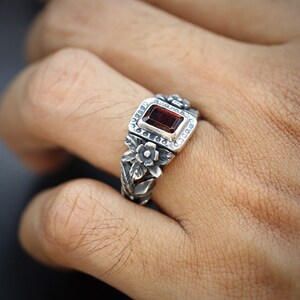 Puede incluir: Un anillo de plata con una piedra preciosa roja engastada en un marco cuadrado rodeado de detalles florales.