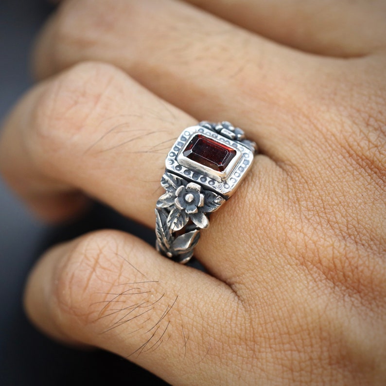 Puede incluir: Un anillo de plata con una piedra preciosa roja engastada en un marco cuadrado rodeado de detalles florales.