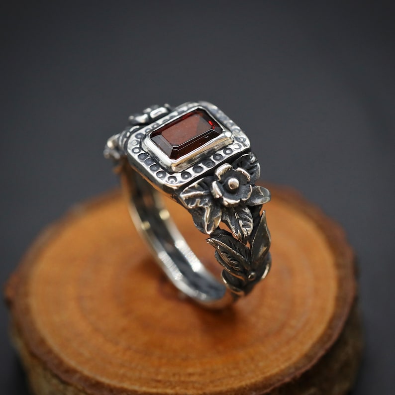 Puede incluir: Un anillo de plata con una piedra preciosa roja y detalles florales. El anillo est&aacute; colocado sobre una superficie de madera.