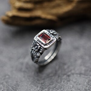 Puede incluir: Un anillo de plata con una piedra preciosa roja engastada en un marco cuadrado. El anillo est&aacute; decorado con detalles florales.