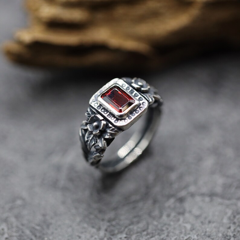 Puede incluir: Un anillo de plata con una piedra preciosa roja engastada en un marco cuadrado. El anillo est&aacute; decorado con detalles florales.