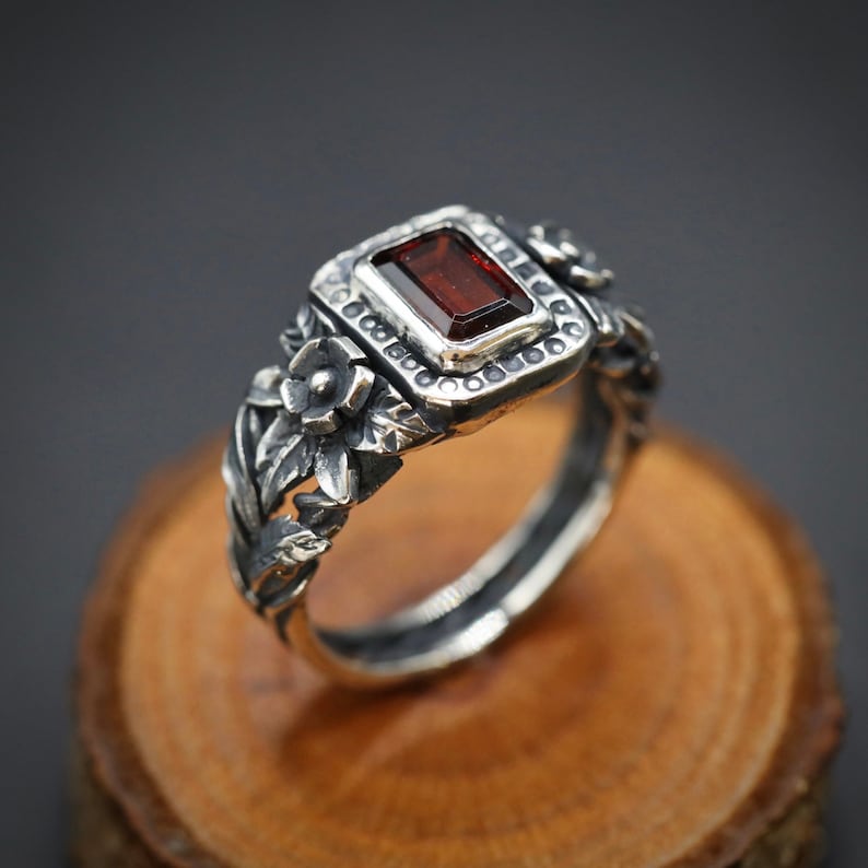 Puede incluir: Un anillo de plata con una piedra preciosa roja engastada en un dise&ntilde;o floral. El anillo est&aacute; sobre una superficie de madera.