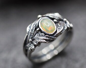 Puede incluir: Un anillo de plata con un delicado diseño de hojas y una piedra de ópalo blanco en el centro.