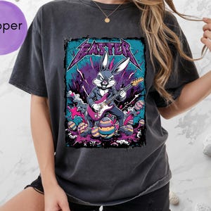 May include: Dark gray t-shirt featuring a graphic of a bunny playing an electric guitar with the word "Easter" in a metal font. The bunny is surrounded by colorful Easter eggs. The shirt has a relaxed fit.